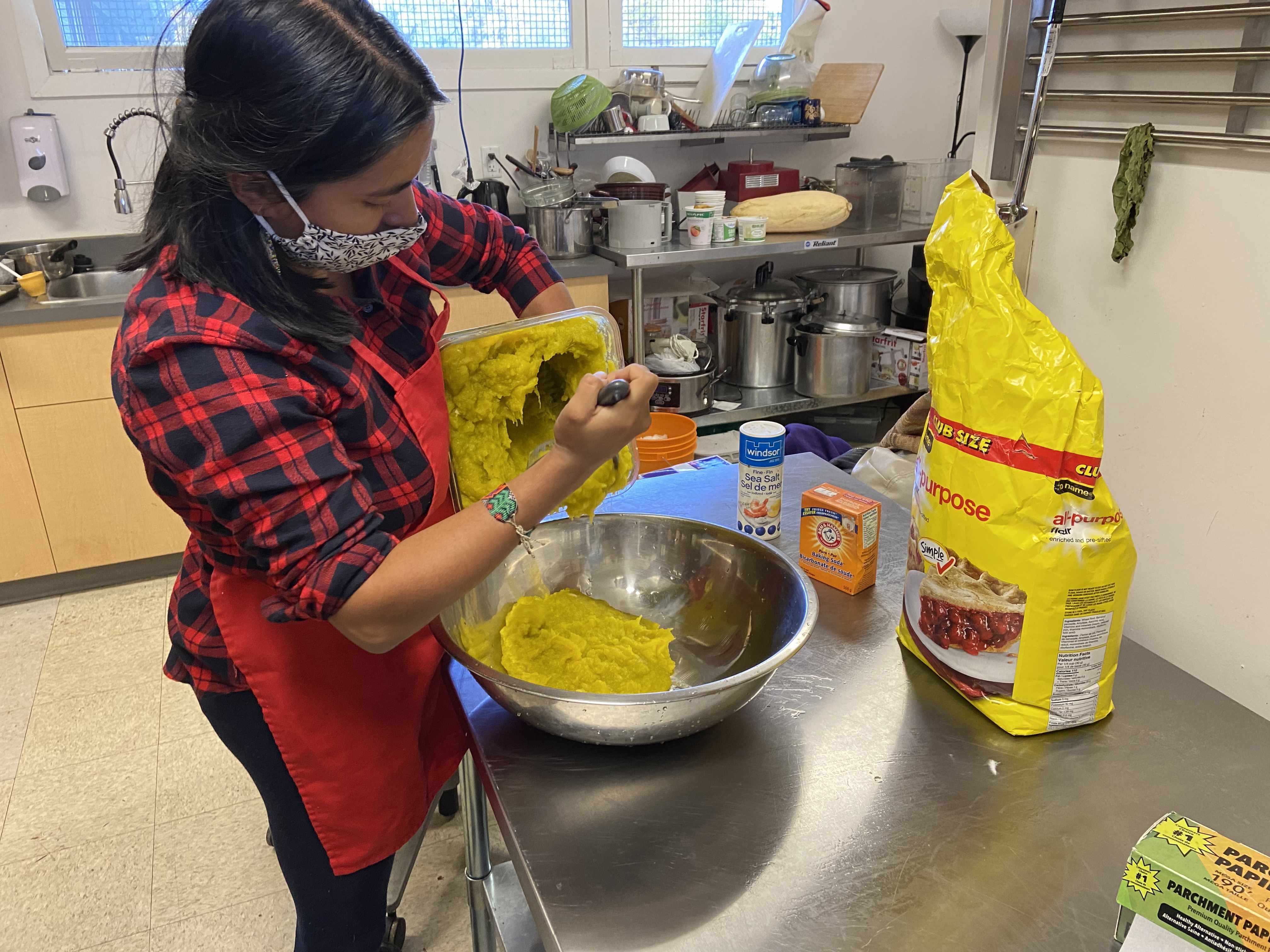 Ingrid mixing a flour and squash mixture into a metal bowl on a countertop alongisde a bag of flour, a box of baking soda and a container of salt
