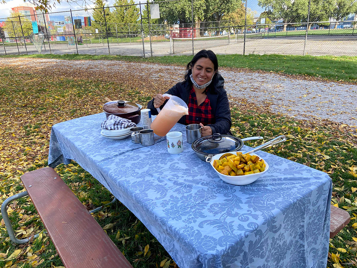 Ingrid sitting at a picnic table covered in a blue floral print, covered in various pots and pans filled with food. She is pouring juice into a cup and smiling