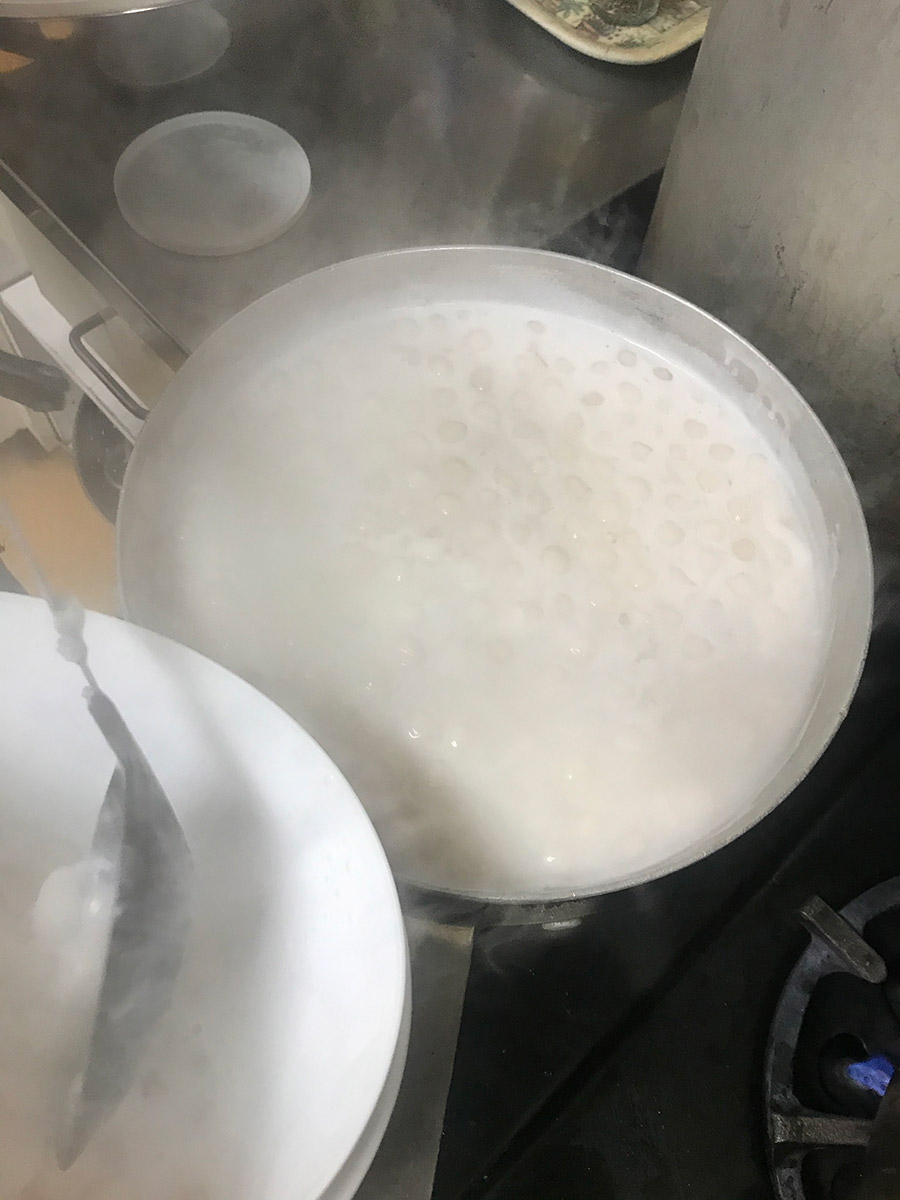 a bowl of what will become a dessert called chè khoai lang, steaming in a saucepan overtop an industrial kitchen stovetop. it is white, thick and creamy with small marble sized homemade tapioca pearls that look like what bubble tea feels like on the tongue
