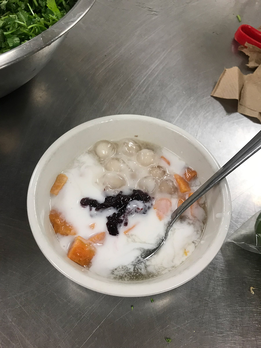 A bowl sitting on a silver vountertop full of chè khoai lang. the bowl contains a thick white sauce, orange cubes of sweet potato with a dollop of mixed berry jam in the center