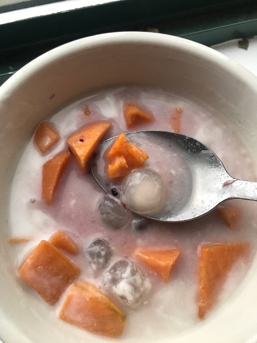 A closeup photograph of a half eaten bowl chè khoai lang with a spoon that contains one sweet potato and one tapioca pearl