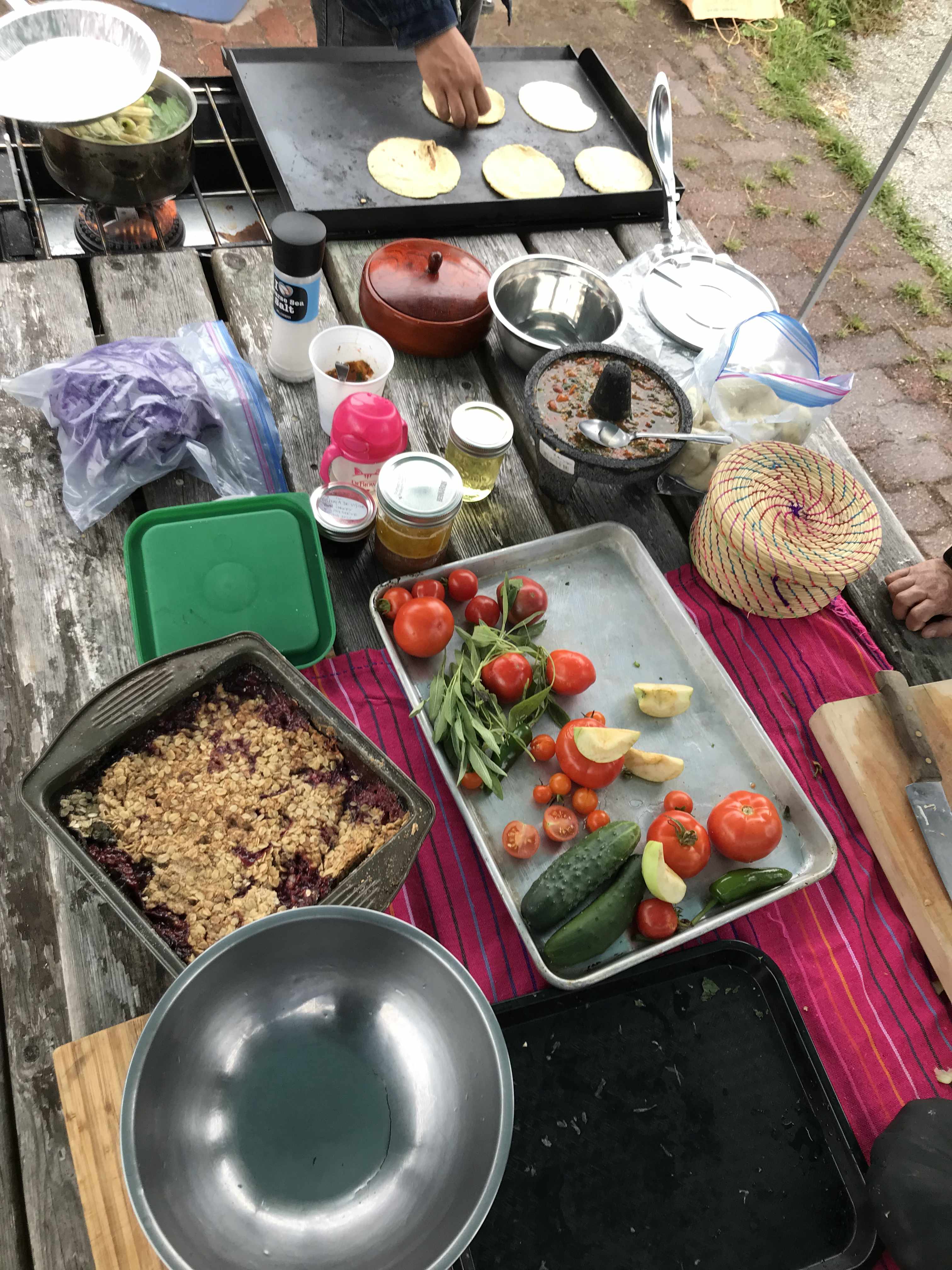 A table top covered with tortillas, salsa, squash, salmon and apple tea. someone is preparing fresh tortillas in the corner of the photo