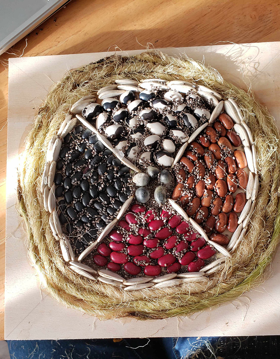 A wheel of various beans placed on a tabletop. the wheel is encased with plant husks and the center is edorned with different beans divided into four quadrants. Each quadrant contains a different variety of beans