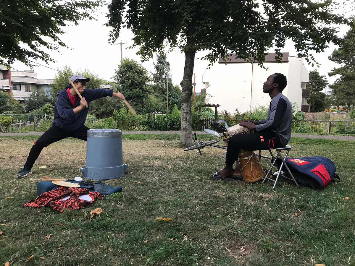 Kage drumming on an overturned garbage can standing across from moses sitting while drumming