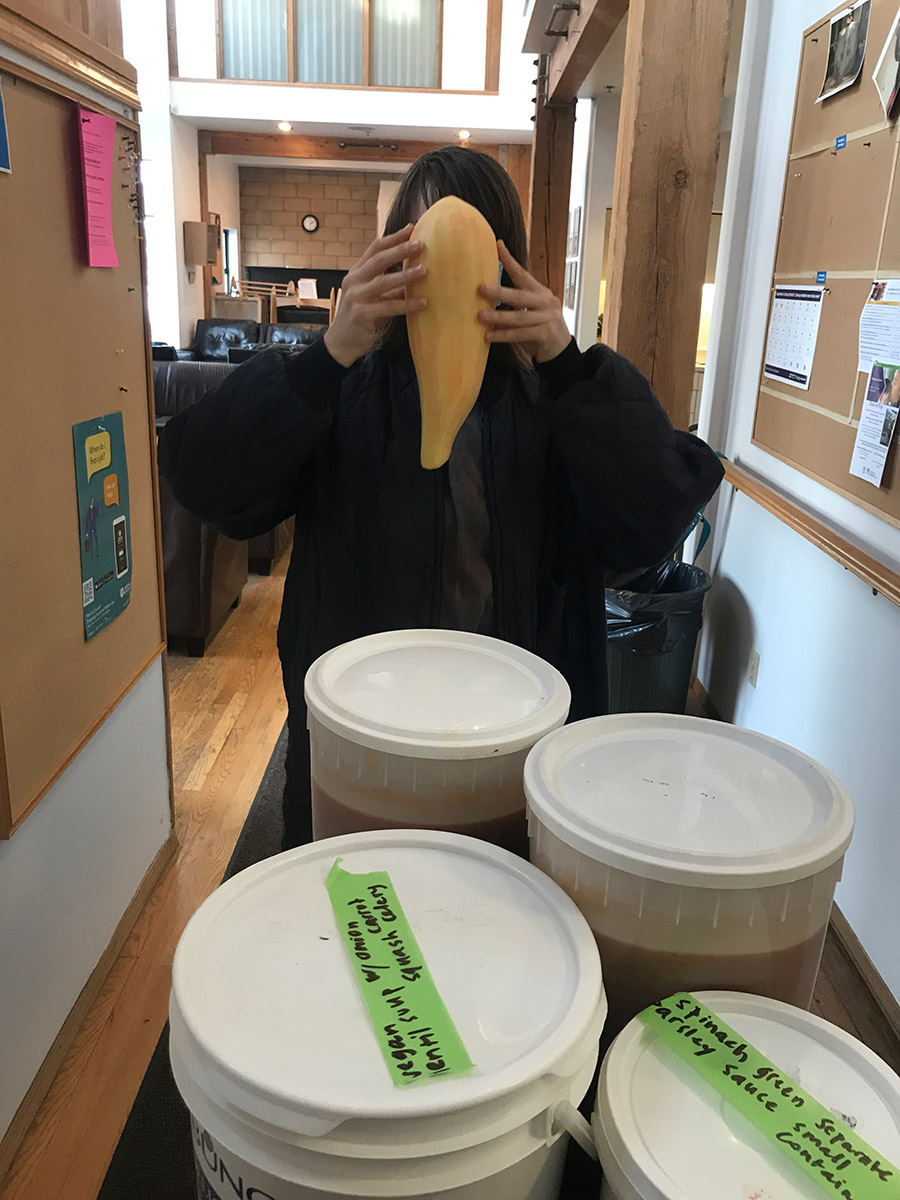 four 20 liters icecream buckets full of soup on a cart. behind the cart is Vivienna in a long sleeve jackt and their face is hidden behind a peeled squash.