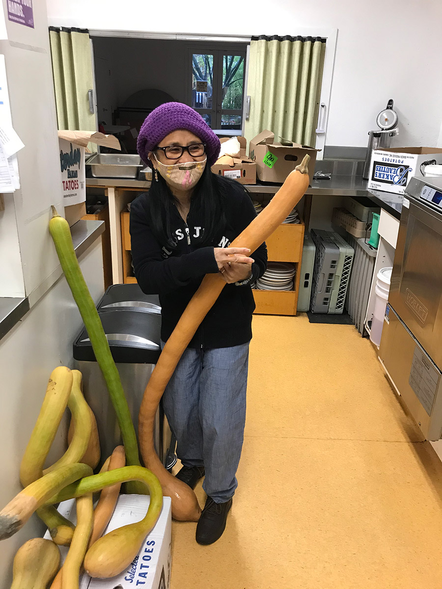 a small framed elderly person with a big smile hidden behind a protective face mask laughing while holding a tromboncino squash as tall as her. there are more squashes on her right side. in the background is an industrial dishwasherand with steel countertops. all the surfaces are full of cardboard boxes and produce