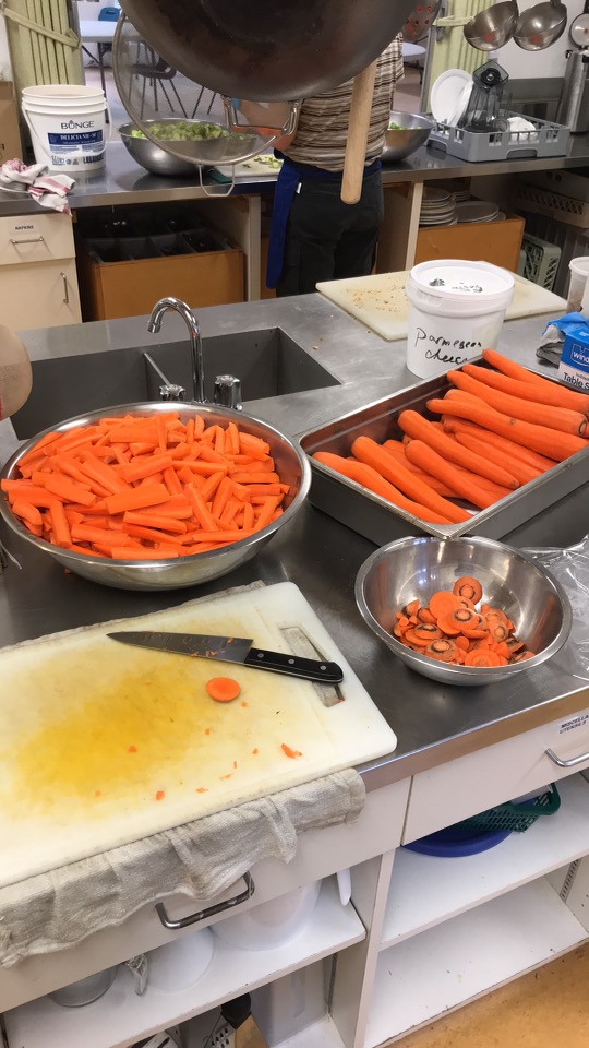 kitchen prepping station of carrots. a sink is in the background. and two bowls and one hotel pan. there are washed and peeled carrots in one bowl, carrot tops in another and quartered 4 inch long carrots in one bowl. a knfe and cutting board in the foreground. total of 20 kilograms or so of carrots.