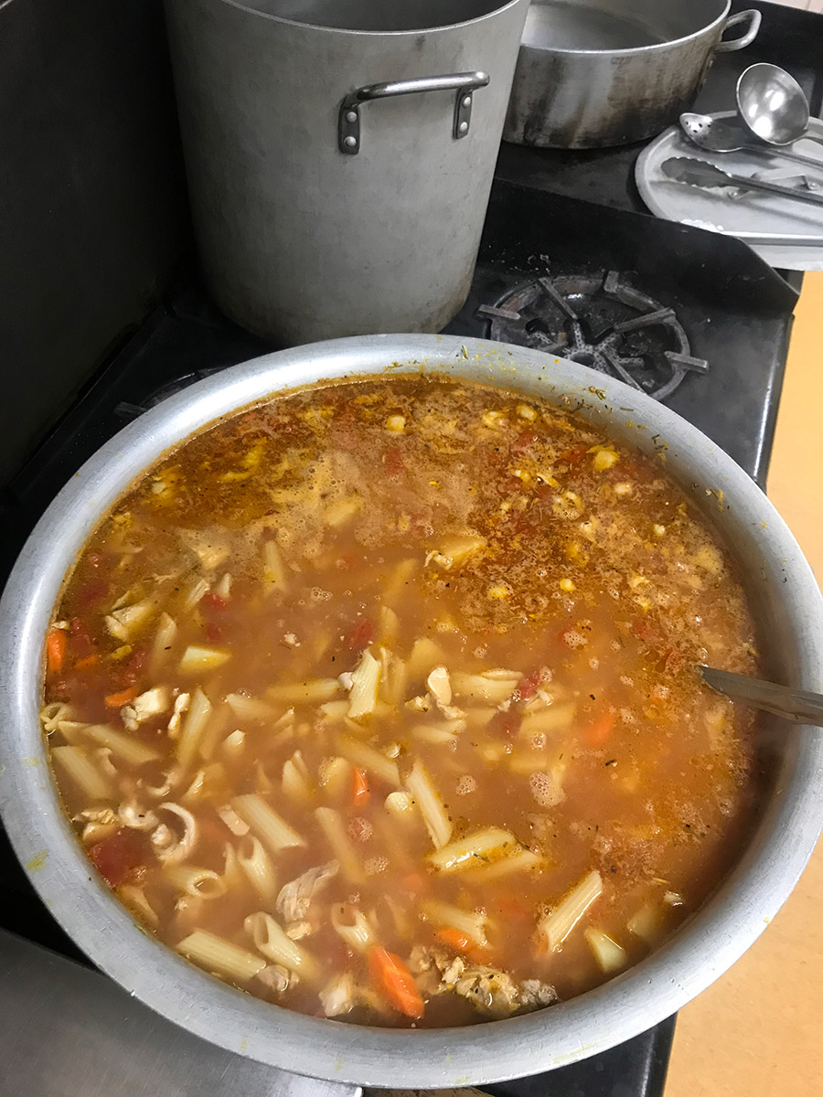 40 litre pot of soup with floating chicken, penne pasta, tomatoes, carrots and oil floating. the pot is full to the brim.