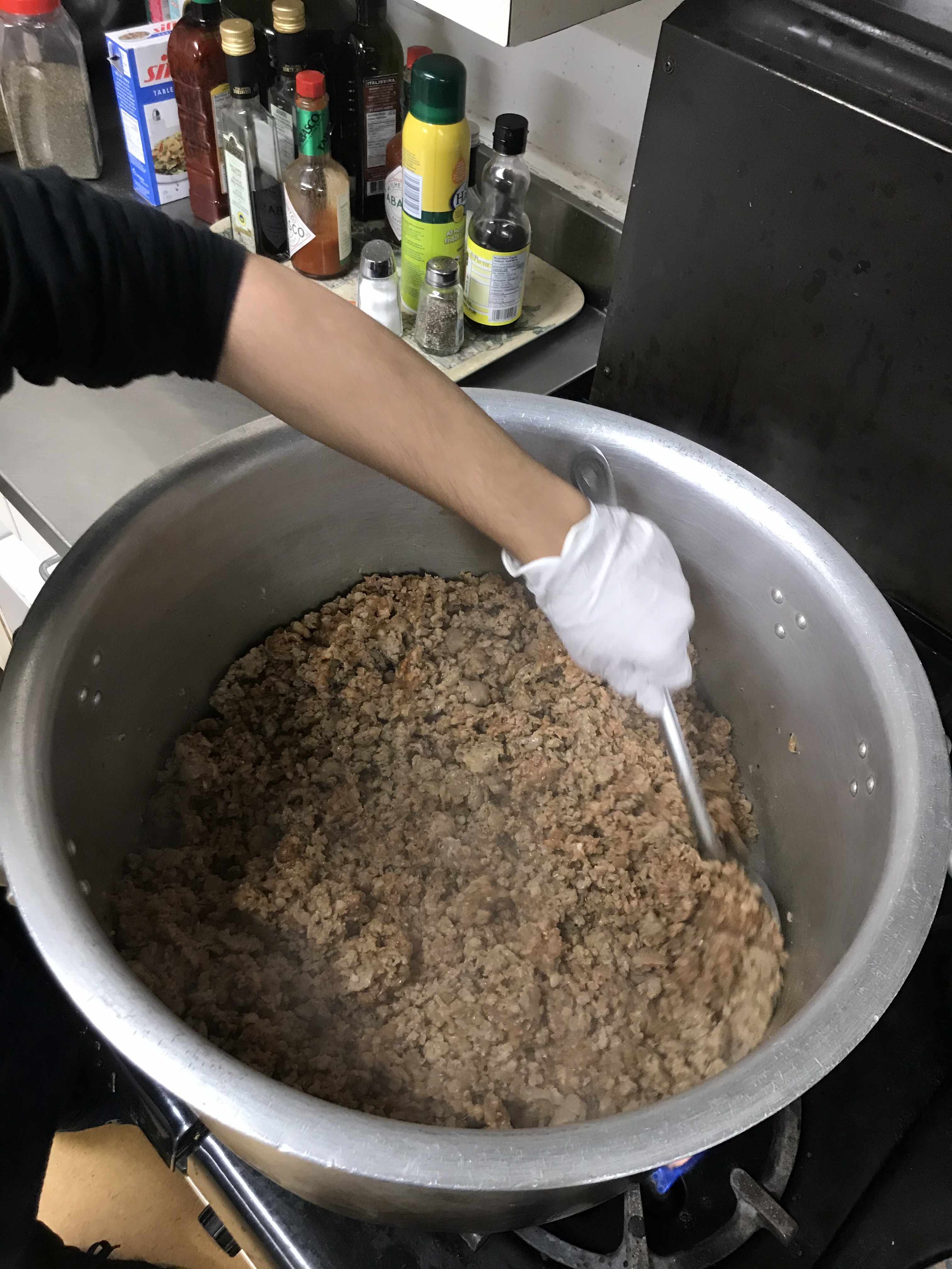 A 40 litre pot of minced meat, mixed with a metal spoon by a hand with gloves. there are seasoning bottles of oil, salt, hot suace and vinegars in the background