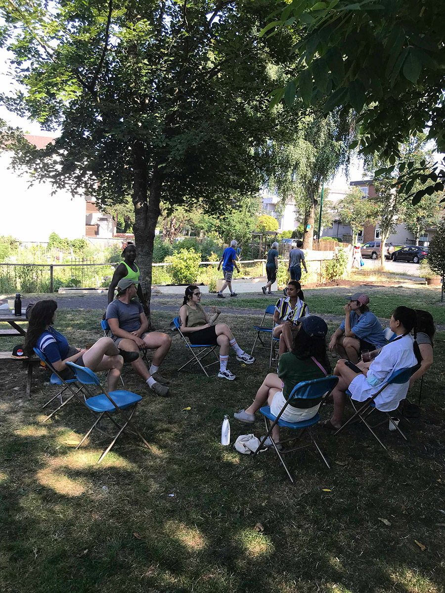 a dozen people seated in a circle outside in the park in daylight, in plastic folding chairs, conversing with food and drinks around them.
