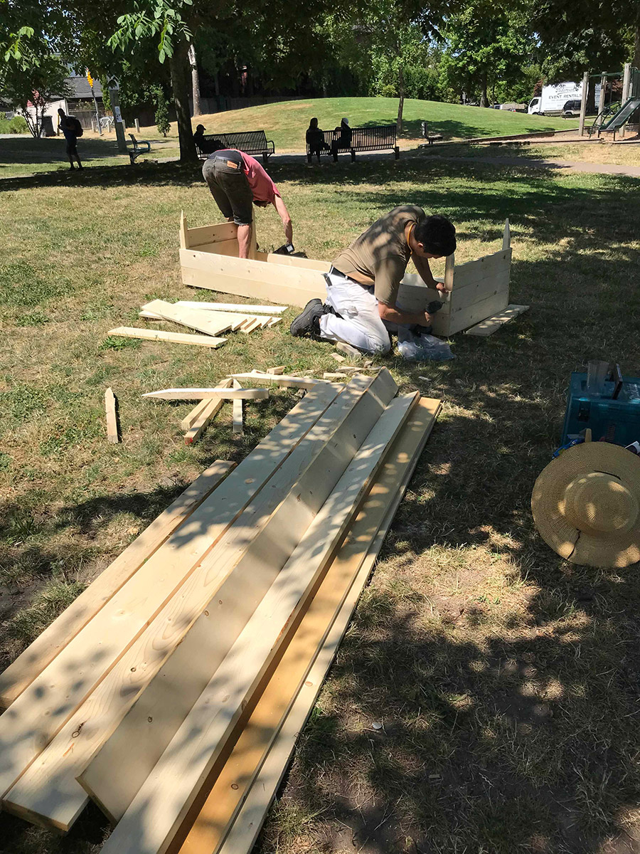 two kneeled figures in the back working on building a raised bed wit drills in their hands. there are lumber scattered around them, and some power tools. they are in the park, working in the sun and shade mixed.
