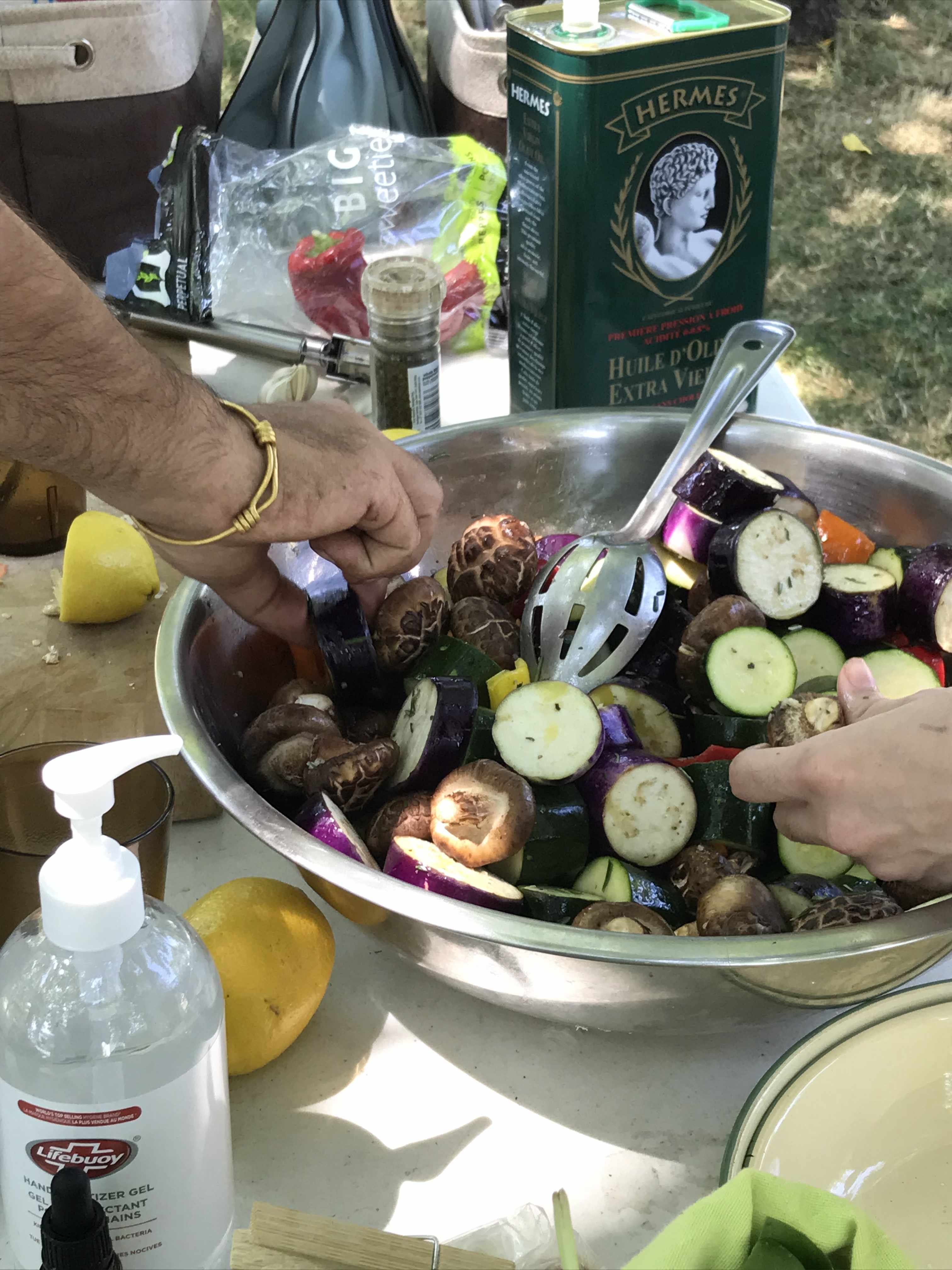 large bowl of marinated vegitables, seasoned for the shish kebabs, on a table. on the table is hand sanitizer, lemons, peppers, black pepper, a tin of olive oil, and two hands are reaching into the bowl to take the fruit.