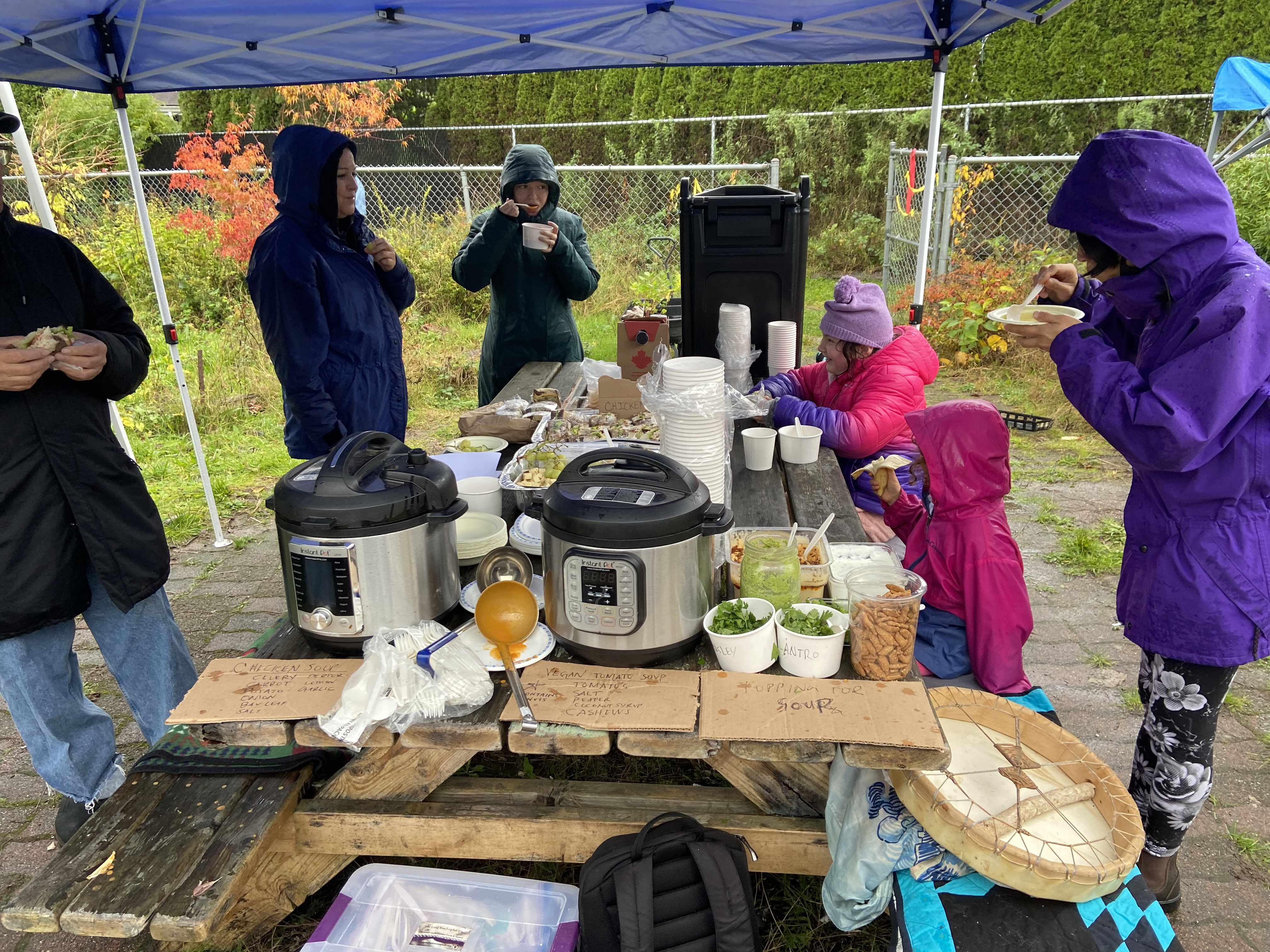 group of 6 people huddled around a picnic bench outdoors in the rain, under a tent. there are lots of cups and food and dirty ladles and everyone is eating and drinking tea