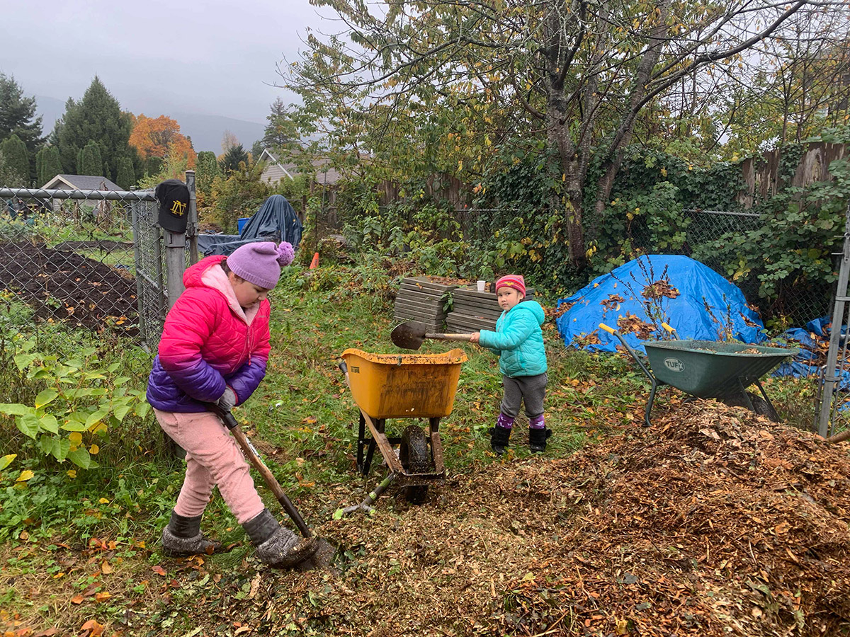 an elementary school age and a kindergarten school age child shovelling mulch into a wheelbarrow