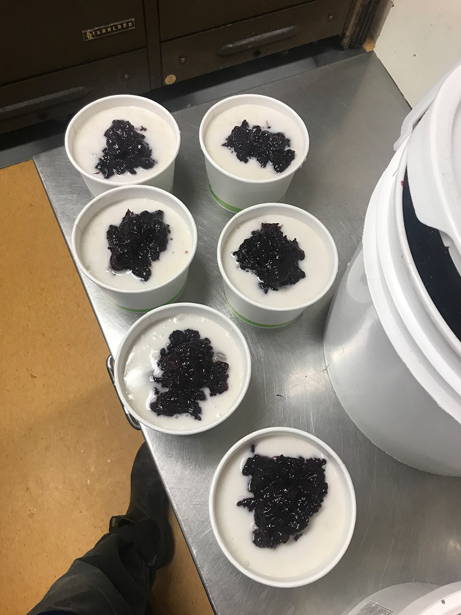 A silver countertop with six containers of chè khoai lang beside a large container of mixed berry jam, viewed from above. A shoe can be seen in the lower left corner