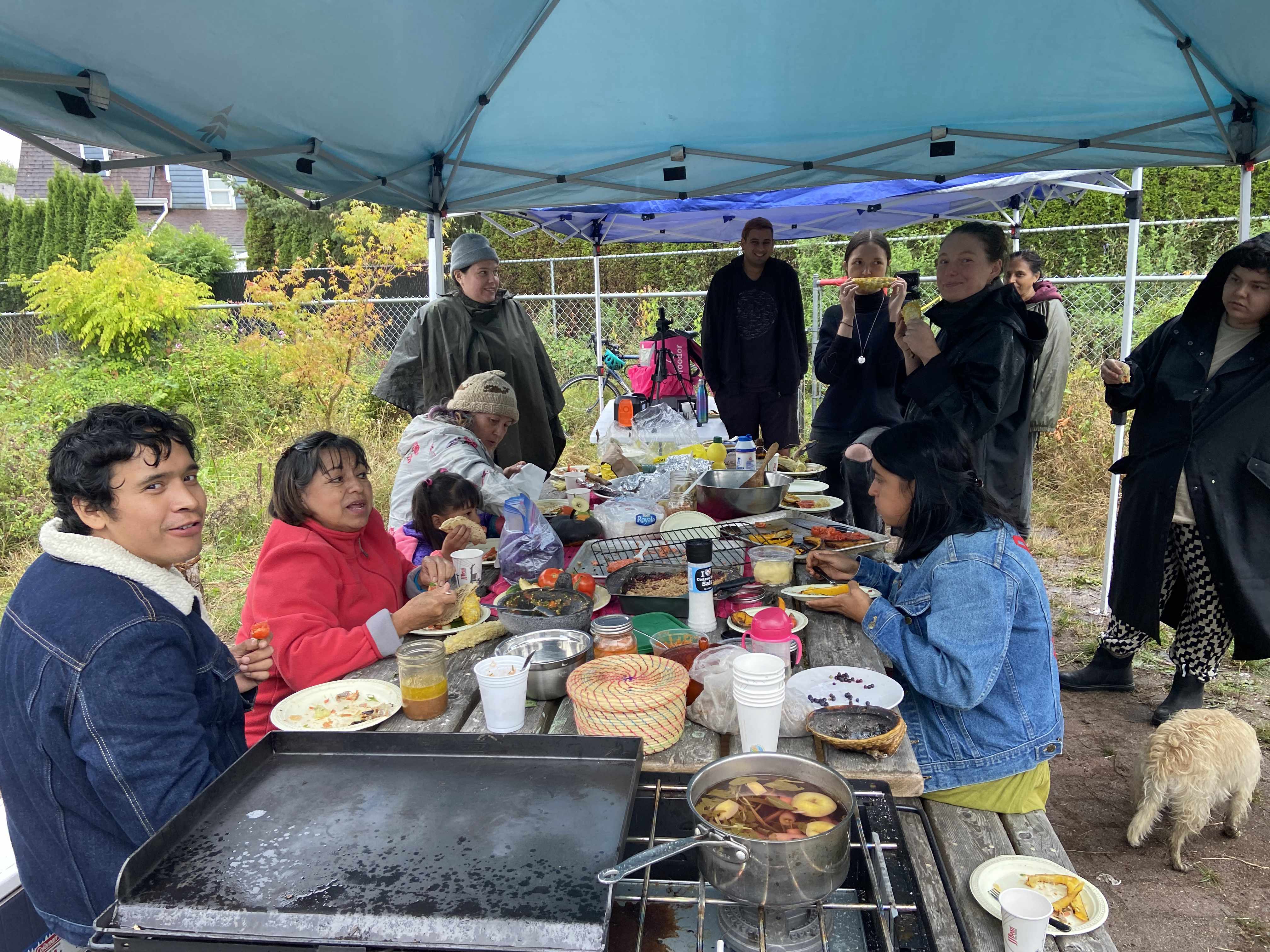 Gardeners and guest at harmony garden sitting around the picnic table under a tent smiling and eating the feast. Beside the table a dog faces aay from the camera.