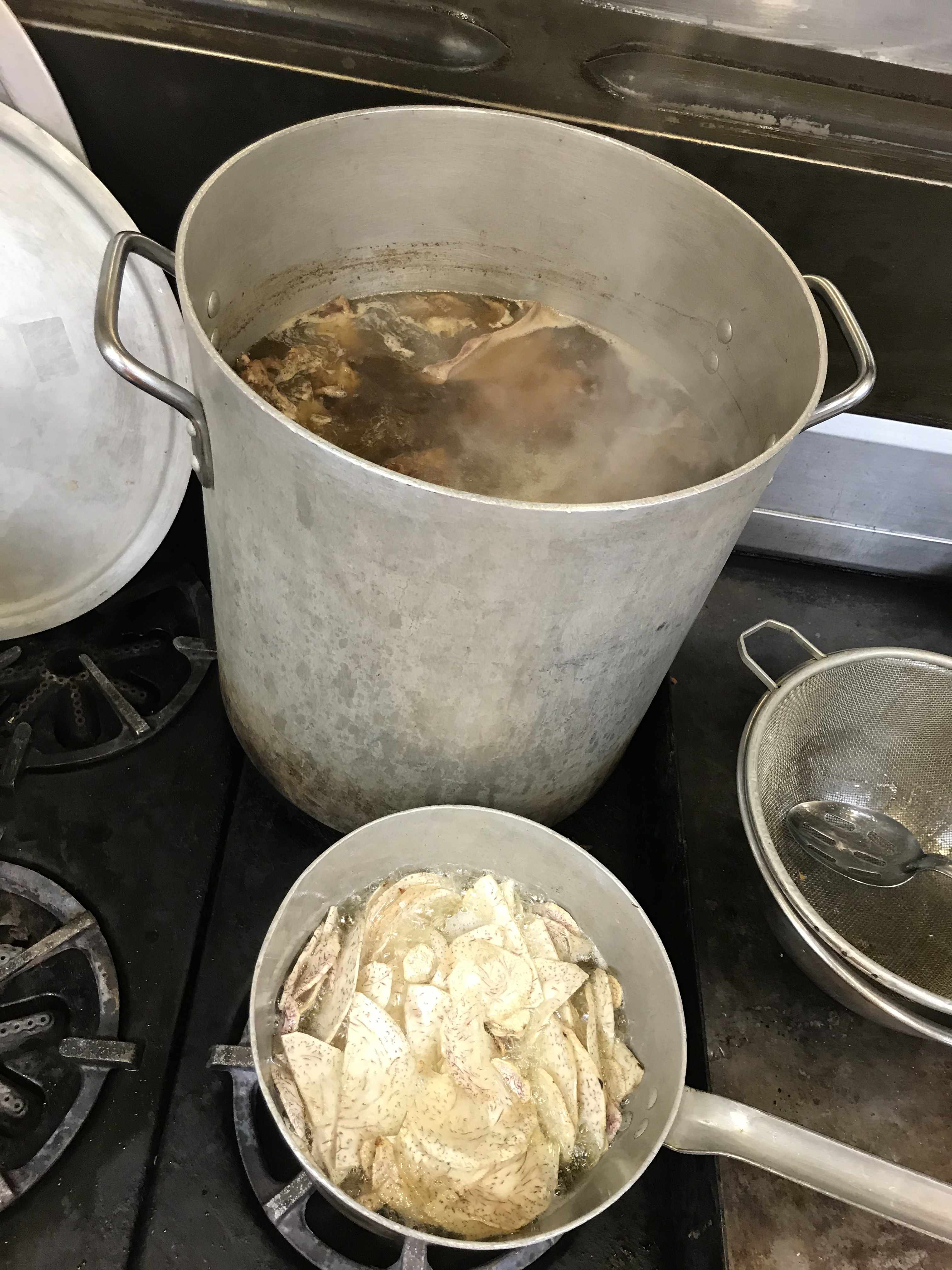 a 40 Litre stockpot on the stove steaming with bone broth and a small saucepan with taro chipps inside being deep fried