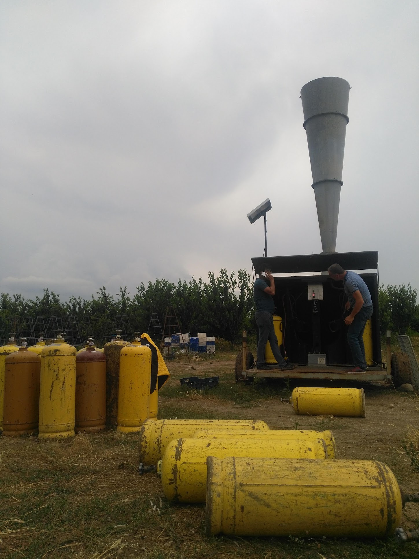 two figures on a box looking machine with a 20 foot inverted cone shape reacing towards the sky. there are a dozen large gas tanks around the figures who seem to be installing a pair of the tanks to the cone machine.