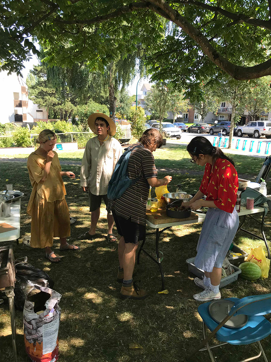 four figures standing around a foldable table talking and working at prepairing lunch. it is sunny and they are under a tree. there is a watermelon on the ground and a bag of charcoal.