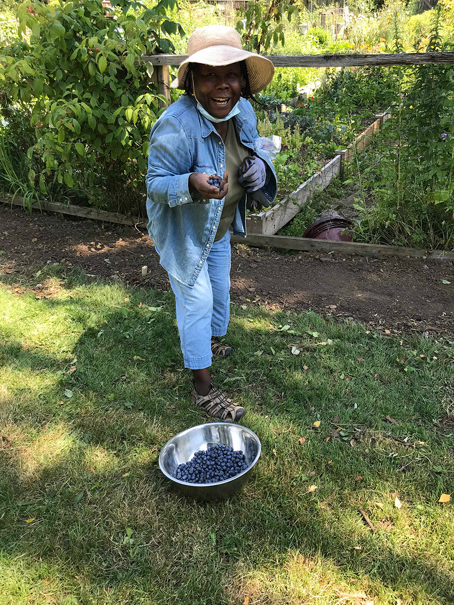 adassa, standing in front of the sahalli garden fence, with a handful of blueberries and a huge smile looking at the camera. she has gardening gloves and tools in her other hand.