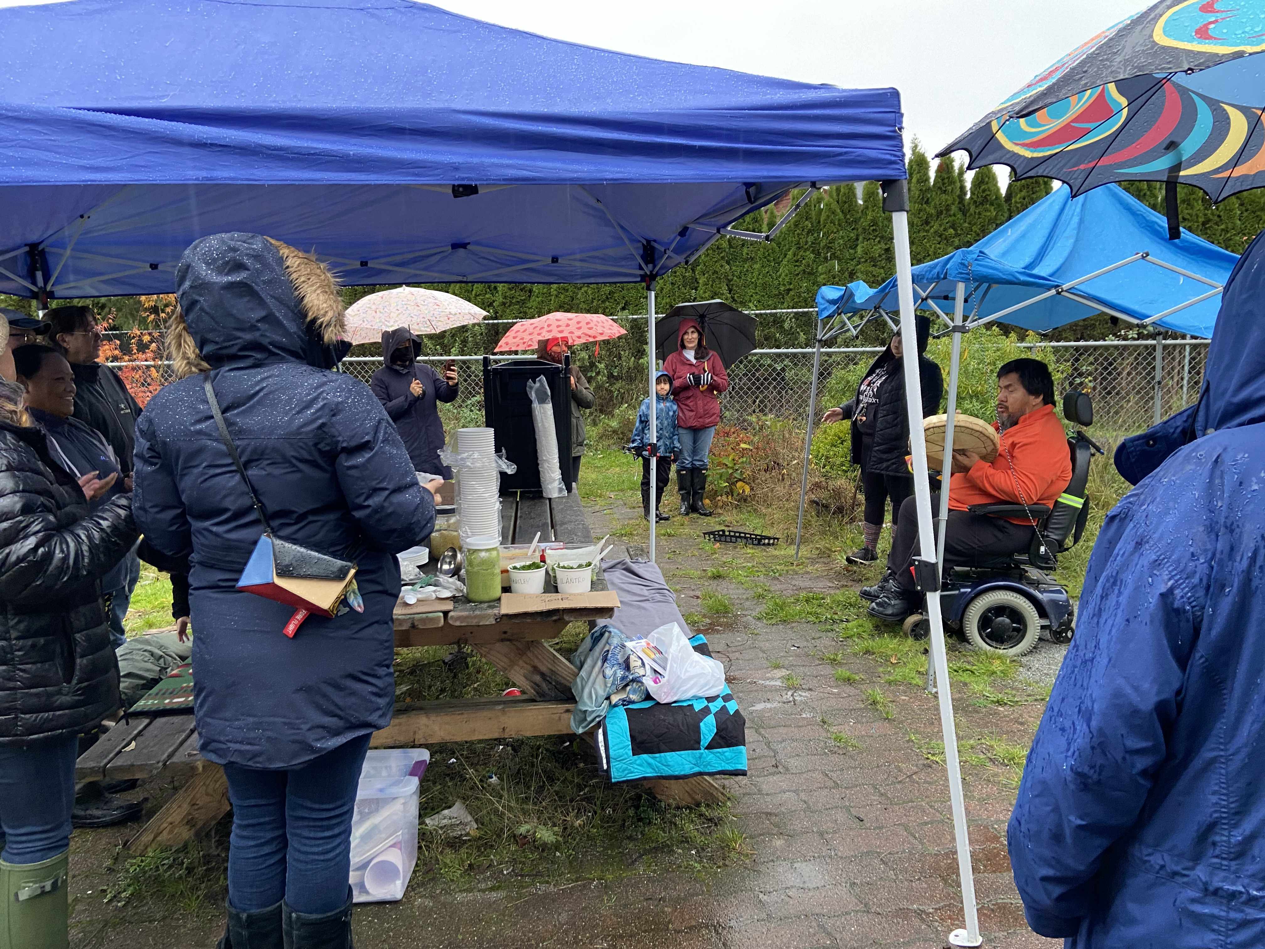 a dozen people standing outside in the rain under tents and umberallas. everyone is listening to the welcome song by Frances who is seated on a motorized chair in an orange shirt with a drum in his hand.