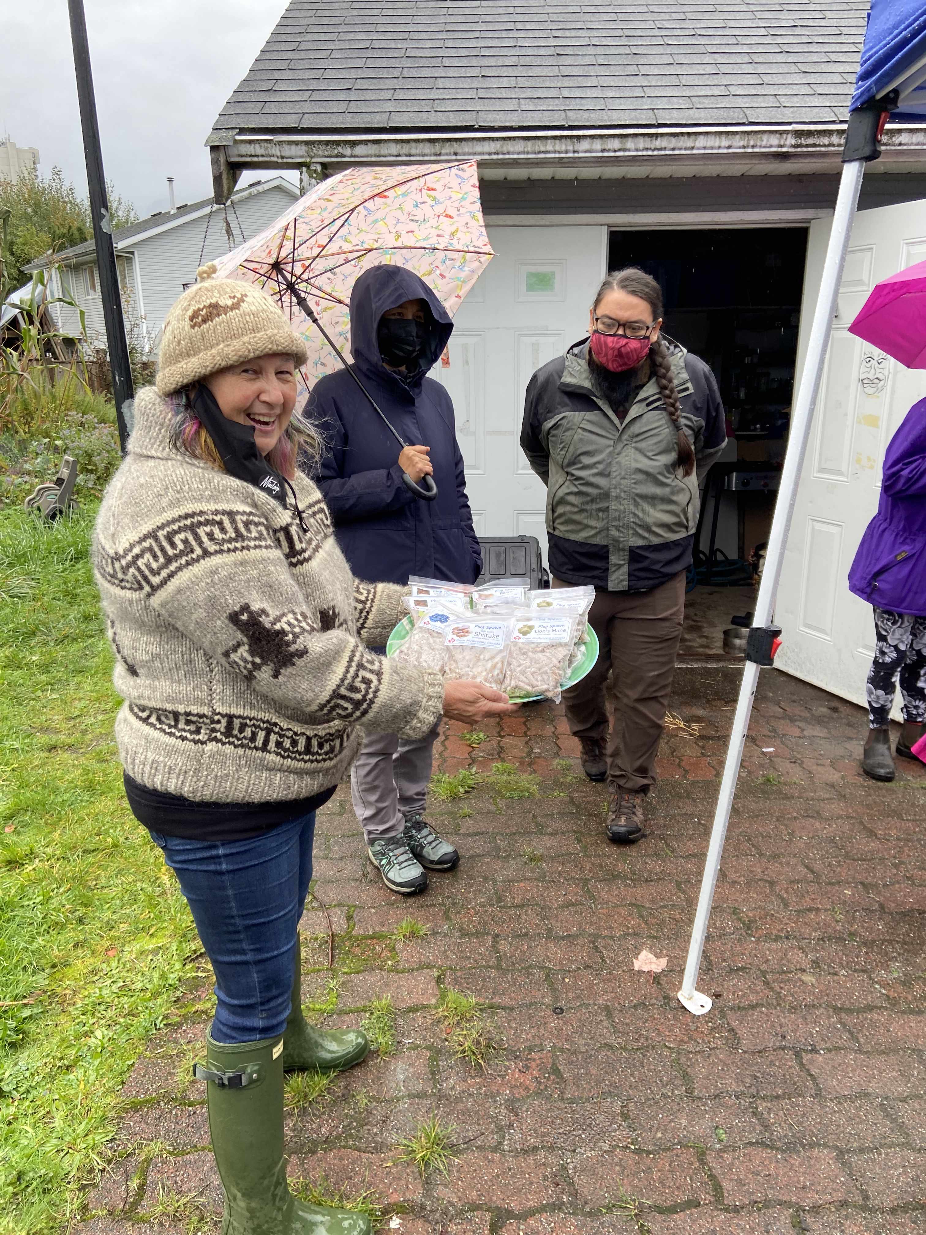 three figures standing in the rain, cease wyss in a cowichan sweater and rubber boots in the foreground holding a tray of mushroom growing kits with a big smile on her face. everyone is standing outside on a brick path in a garden