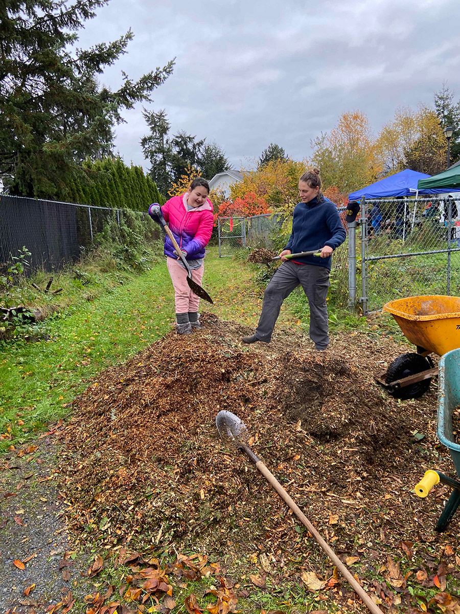 an elementary school aged child and adult on a pile of garden mulch, shovelling the mulch into wheelbarrows